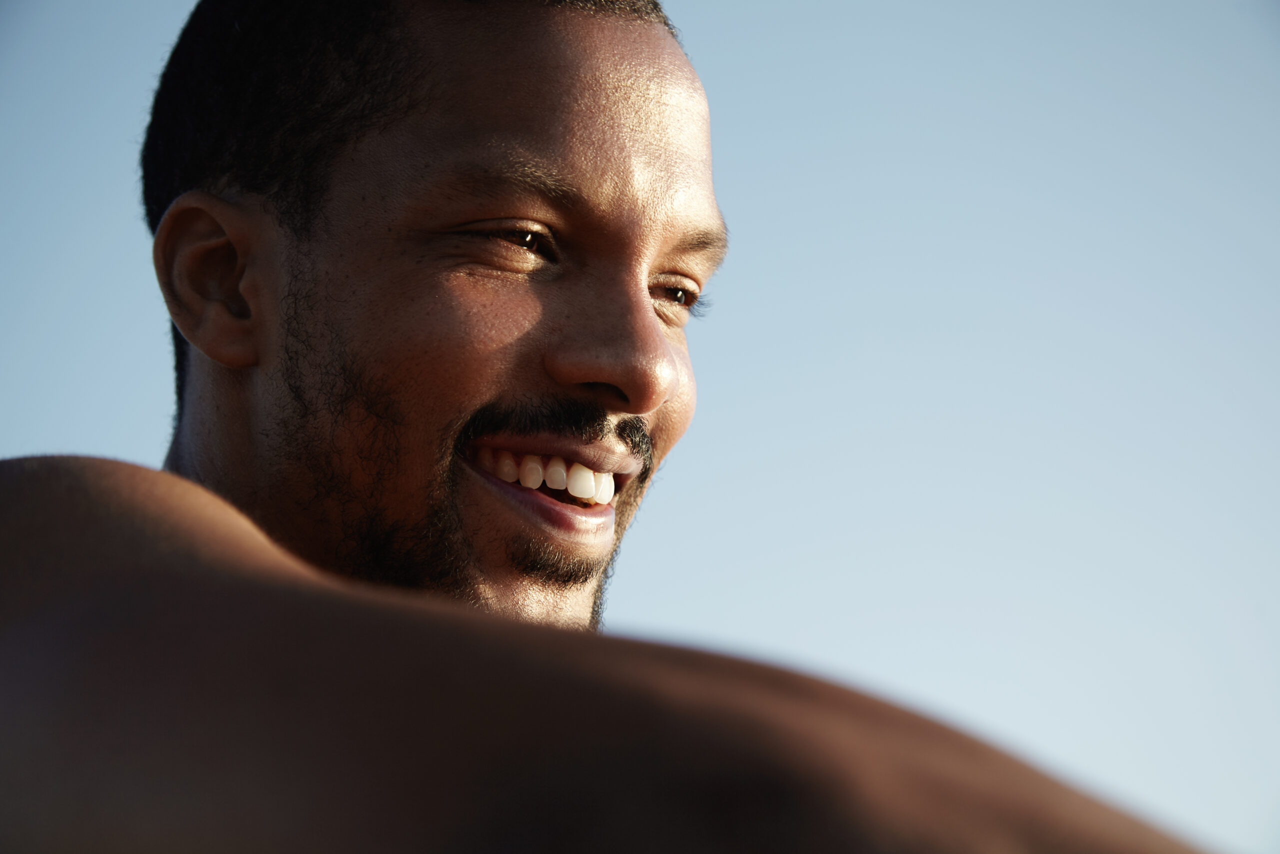 close up view of handsome happy dark skinned man with healthy skin and cheerful smile looking into distance, squinting his eyes while relaxing alone outdoors, enjoying sun against blue sky background